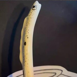 May include: A ceramic eel figurine with a cream-colored body and black spots. The eel has a large, round eye and a curved, upward-facing posture. The figurine is set against a dark background.