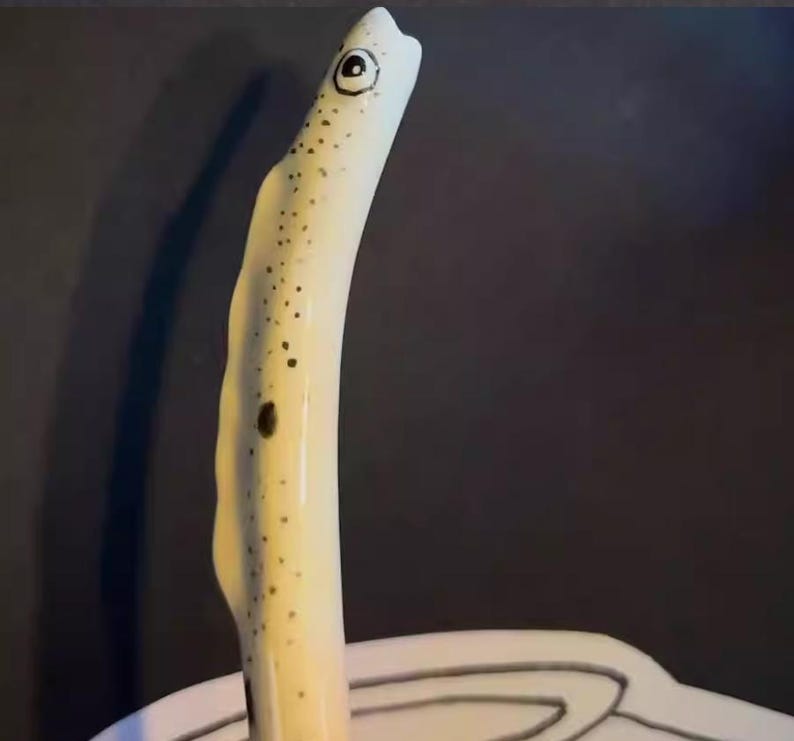 May include: A ceramic eel figurine with a cream-colored body and black spots. The eel has a large, round eye and a curved, upward-facing posture. The figurine is set against a dark background.