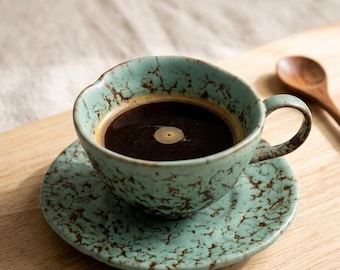 May include: A teal ceramic coffee cup and saucer set filled with dark coffee. The cup and saucer have a speckled, crackle glaze design. A wooden spoon rests on a wooden surface next to the cup.