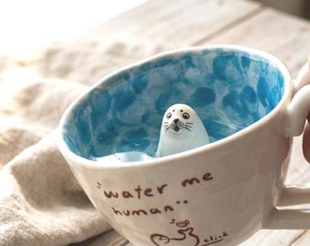 Puede incluir: Una taza de cerámica blanca con interior jaspeado en azul que presenta una pequeña figura de foca esculpida en el fondo. El exterior es de color blanco roto con el texto marrón "water me human" y un dibujo de una foca con la palabra "click".