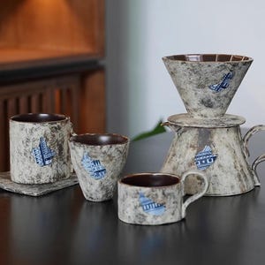 May include: A ceramic coffee set with a textured, speckled finish. The set includes a pour-over coffee maker, a mug with a handle, two cups, and a small tray. Each piece features a blue geometric design.