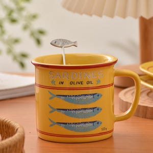 Könnte beinhalten: Gelbe Keramik-Tasse mit rotem Rand und Henkel, mit Sardinen-Illustrationen und dem Text "Sardines in Olive Oil". Ein kleiner löffel in Fischform liegt in der Tasse. Die Tasse ist 125g.