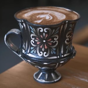 May include: A dark ceramic mug with a handle, filled with a latte. The mug features a floral design in white, pink, and blue against a black background. The latte art is a heart shape.