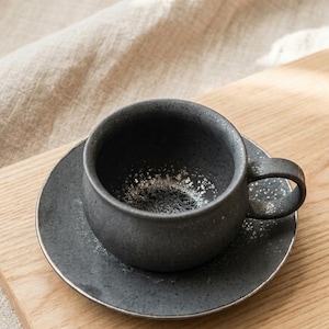 Könnte beinhalten: Ein dunkelgraues Keramik-Tee-Tassen- und Untertassen-Set. Die Tasse hat eine abgerundete Form mit einem Henkel, und die Untertasse ist kreisförmig. Beide Teile haben eine gesprenkelte Textur und einen silbernen Rand. Das Set steht auf einer Holzoberfläche.