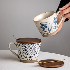 Puede incluir: Tazas de cerámica con diseños florales azules. Una taza tiene una tapa de madera y una cuchara dorada. La otra taza se levanta. Las tazas tienen un fondo beige con diseños florales azules y bases marrones.