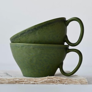 Puede incluir: Dos tazas de cerámica verde oliva apiladas con asas redondeadas. Las tazas tienen una superficie texturizada y una forma ligeramente acampanada. Están sobre una tela beige texturizada.