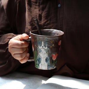 Puede incluir: Una taza de cerámica marrón con asa, con una ilustración pintada a mano de un perro. El perro está representado en tonos grises y blancos, rodeado de tulipanes rojos y follaje verde. La taza también tiene una cuchara a juego.