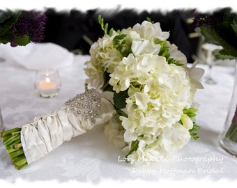 May include: A white bridal bouquet with white hydrangeas and freesia, wrapped in white satin ribbon with a jeweled embellishment. The bouquet is on a white tablecloth with a candle in the background.