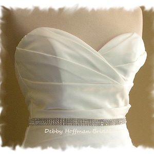 May include: A white strapless wedding dress with a crystal rhinestone belt. The belt is thin and has a delicate sparkle. The dress is made of a smooth, satin fabric.