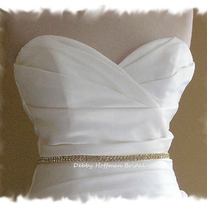 May include: A white strapless wedding dress with a simple, elegant design. A thin, sparkly gold belt with rhinestones is worn around the waist. The text "Debby Hoffman Bridal" is visible below the belt.