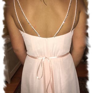 May include: A light pink chiffon bridesmaid dress with a low back featuring a double strap with rhinestones. The dress has a satin sash tied in a bow at the back.