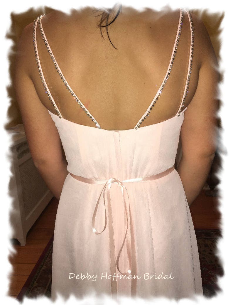 May include: A light pink chiffon bridesmaid dress with a low back featuring a double strap with rhinestones. The dress has a satin sash tied in a bow at the back.