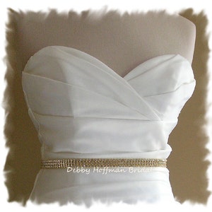 May include: A white strapless wedding dress with a gold rhinestone belt. The belt has the text "Debby Hoffman Bridal" on it.