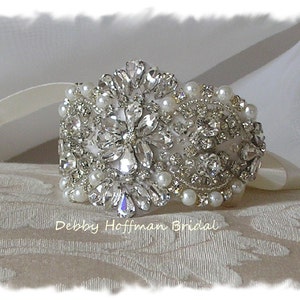 May include: A silver and white bridal bracelet with a large crystal and pearl design. The bracelet is on a white fabric background.