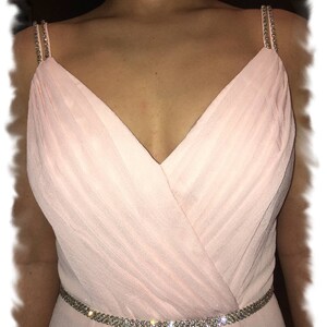 May include: A blush pink chiffon bridesmaid dress with a V-neckline and a crystal embellished belt. The dress has thin straps with crystal embellishments. The dress is labeled "Debby Hoffman Bridal".
