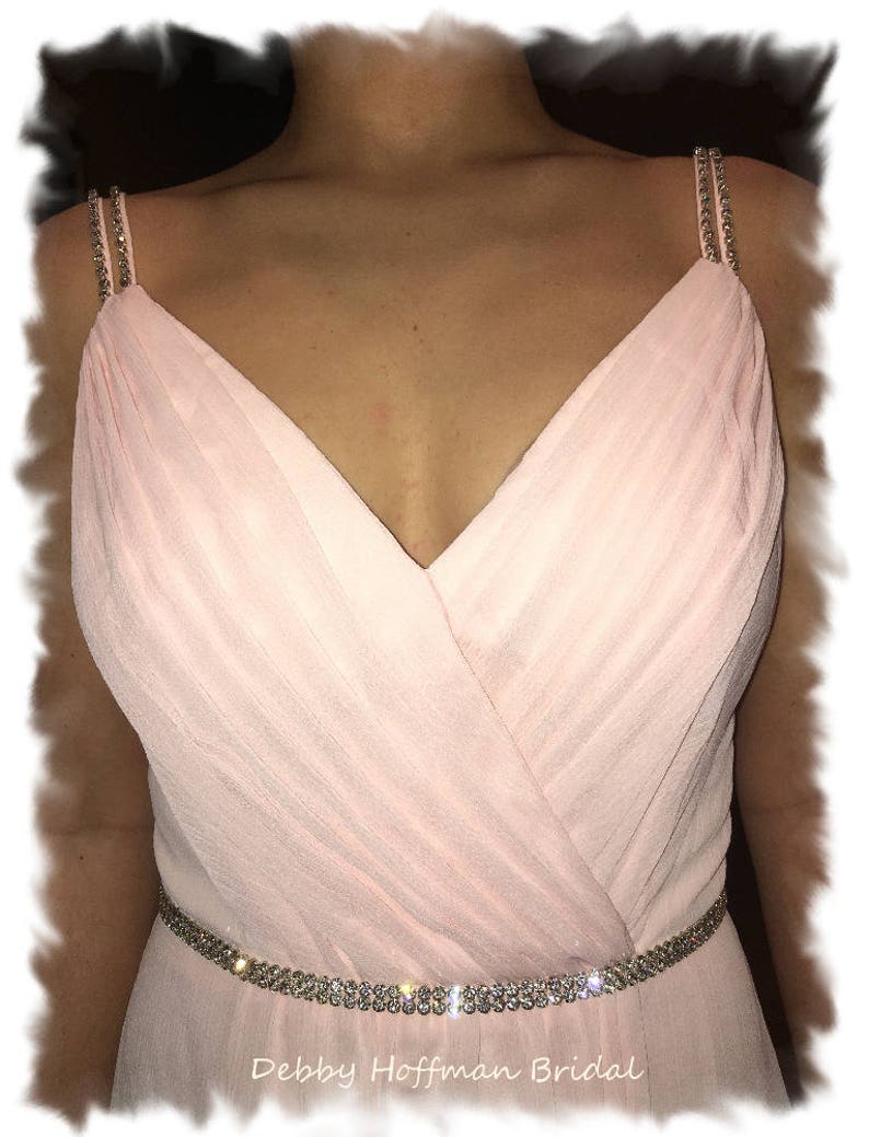 May include: A blush pink chiffon bridesmaid dress with a V-neckline and a crystal embellished belt. The dress has thin straps with crystal embellishments. The dress is labeled "Debby Hoffman Bridal".