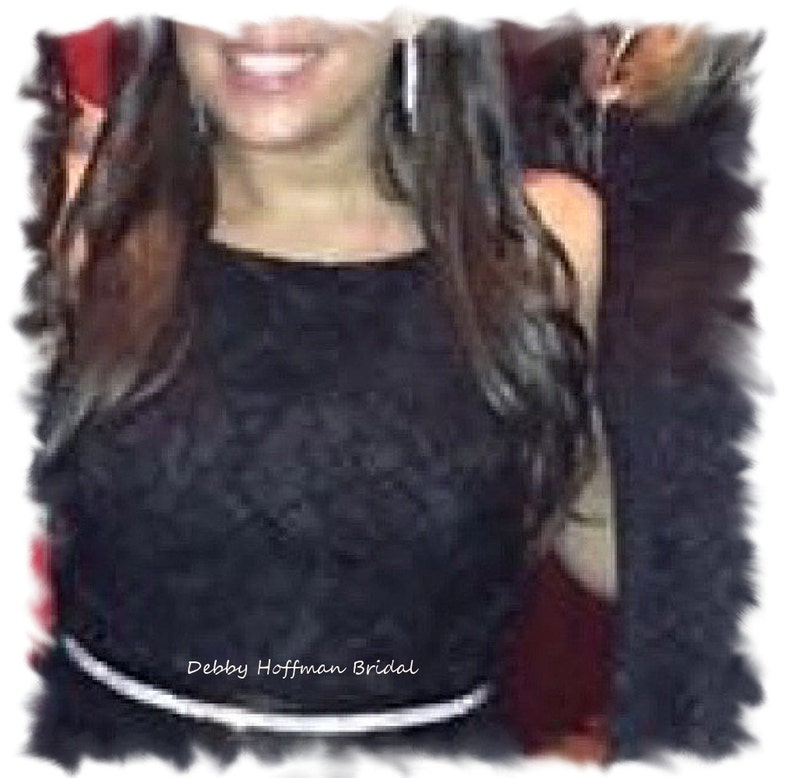 May include: A woman wearing a black lace dress with a white belt. The dress has a scoop neckline and a fitted bodice. The woman is smiling and has long brown hair. The text "Debby Hoffman Bridal" is visible on the dress.