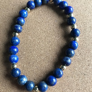Könnte beinhalten: Ein blaues Lapislazuli-Armband mit goldenen Akzenten. Das Armband besteht aus runden Perlen und ist auf einer elastischen Schnur aufgefädelt.