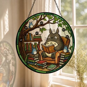 Pode incluir: Vitral redondo com uma cena com um grande personagem cinza Totoro lendo um livro, cercado por personagens azuis menores e livros. O design inclui uma &aacute;rvore e uma borda verde, pendurada por uma corrente.