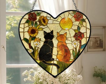 May include: Heart-shaped stained glass window art featuring two cats, one black and one orange, gazing at a yellow sun. The artwork includes sunflowers, roses, and daisies. A black metal chain is attached for hanging. The window is approximately 10 inches wide.