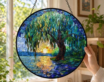 May include: A circular stained glass suncatcher featuring a weeping willow tree beside a body of water at sunset. The design uses shades of blue, green, and yellow glass pieces to depict the tree, the rippling water, and the glowing sun.