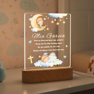 May include: A rectangular acrylic night light with a wooden base featuring a printed illustration of a child on a crescent moon, a sleeping child on clouds, stars, and a cross. The text reads: Mia Garcia, God in heaven hear my prayer.