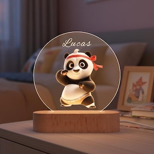 Könnte beinhalten: Dekoratives Nachtlicht mit einem Cartoon-Panda-Bären-Charakter mit rotem Stirnband und einer Kampfsportpose. Der Panda befindet sich vor einem transparenten, kreisförmigen Hintergrund mit dem Namen "Lucas" und steht auf einem Holzsockel.