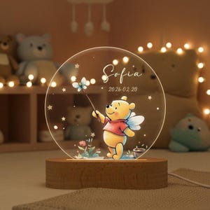 May include: A circular acrylic night light featuring Winnie the Pooh, with the name "Sofia" and the date "2026.02.20". The light has a wooden base and a cartoon illustration of Winnie the Pooh holding a flower. The light is designed for a child's room.