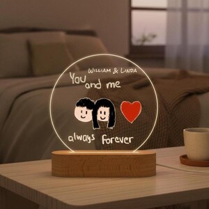 May include: A circular acrylic night light with a wooden base. The light displays the text "You and me always forever" and the names "William & Linda". Two cartoon figures and a red heart are also shown.