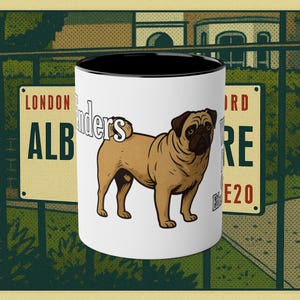 May include: White ceramic mug with a black interior featuring a tan pug illustration. The mug has the word "Pug" in a stylized font. The background includes signs that say "London" and "Ford" with the letters "Alb" and "Re" visible.
