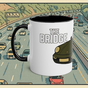 Könnte beinhalten: Weißer Keramikbecher mit schwarzem Innenraum und Henkel. Der Becher zeigt den Text "THE BRIDGE" und eine grüne Autografik. Der Hintergrund zeigt eine Autobahnszene mit Autos und einer Palme.