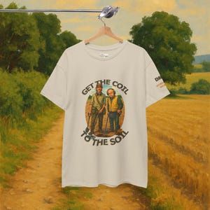 Può includere: T-shirt color crema con grafica di due persone con metal detector. Il testo "GET THE COIL TO THE SOIL" è stampato sopra e sotto l'illustrazione. La maglietta è appesa a una gruccia di legno.