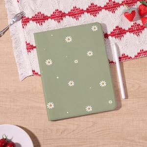 May include: A sage green tablet case adorned with white daisy flower designs and small white dots. A white stylus rests beside the case. The case is on a wooden surface with a red and white patterned cloth.