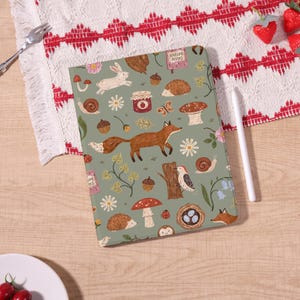 May include: A green tablet case with a whimsical woodland pattern featuring a fox, rabbit, hedgehog, mushrooms, and the words "Nature Notes". The case is on a wooden surface with a white stylus and a red and white patterned cloth.