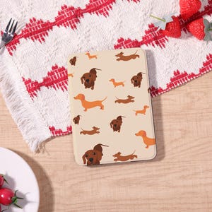 Peut inclure: Une coque de tablette beige avec un motif répété de teckels de dessin animé en marron et orange. L'étui est rectangulaire et a une surface lisse. L'arrière-plan comprend un tissu à motifs blanc et rouge.