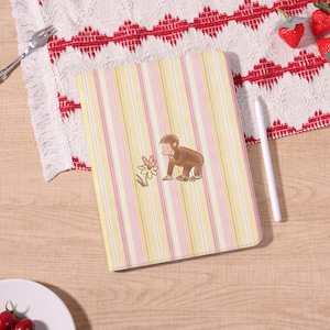 May include: A rectangular notebook with a yellow and pink striped pattern. A brown monkey illustration is centered on the cover, looking at a flower. The notebook is on a wooden surface, with a white stylus and a red and white patterned cloth.