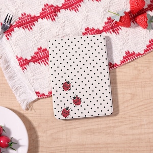 May include: A white tablet case with a black polka dot pattern and three red ladybug illustrations. The case is on a wooden surface next to a white and red patterned cloth.