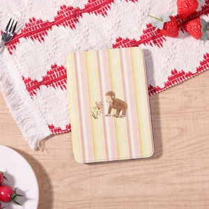 May include: A rectangular notebook with a yellow and pink striped pattern. The cover features a cartoon illustration of a brown monkey and a flower. The notebook is on a wooden surface with a red and white patterned cloth.