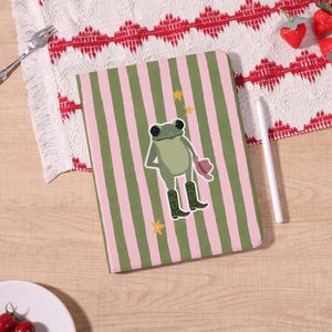 Könnte beinhalten: Ein Notizbuch mit grünem und rosa Streifenmuster. Das Cover zeigt einen Cartoon-Frosch mit Cowboyhut und Stiefeln, mit gelben Sternen. Das Notizbuch liegt auf einer Holzoberfläche mit einem dekorativen Tuch und einem Stift.