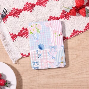 May include: A decorative rectangular item with a pastel floral and bow design. The design includes pink and blue flowers, bows, and hearts on a light blue and pink striped and checkered background. The item is on a wooden surface.