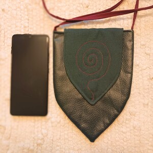 May include: A dark green leather crossbody bag with a flap closure. The flap has a green fabric insert with a red embroidered spiral design.
