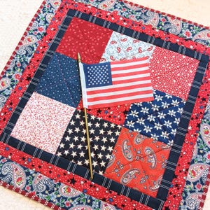 May include: A patriotic quilted placemat featuring red, white, and blue fabrics. The square design incorporates various patterns, including stars, paisley, and solid colors. An American flag is placed on top, adding to the theme.