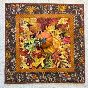May include: A square fabric wall hanging featuring a central panel of autumn leaves, pumpkins, and berries in shades of orange, yellow, and brown. The panel is framed by a mustard yellow border with small dark dots and a dark brown outer border.
