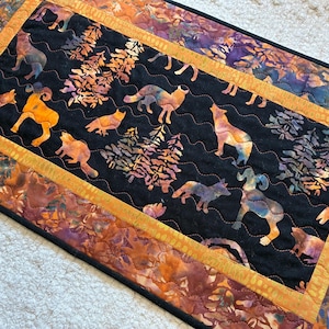 May include: A rectangular fabric table runner featuring a black center panel with a multicolored batik print of wolves, bighorn sheep, and pine trees. The runner has a wavy quilted texture and a wide border in shades of orange and purple.