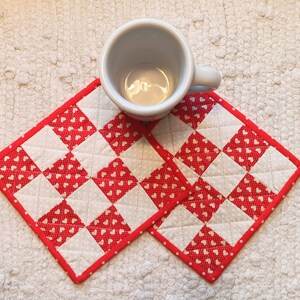 May include: Two red and white quilted fabric coasters with a white heart pattern. A white ceramic mug is sitting on top of the coasters.