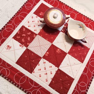 May include: A red and white quilted table runner with a patchwork design. The runner features a variety of patterns, including floral prints and polka dots. A red teapot and teacup with floral accents sit on the runner.