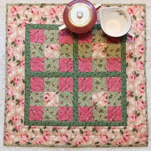 May include: A square quilted placemat with a floral pattern in shades of pink and green. The center features a patchwork design with pink squares and green squares. A red teapot and a white teacup with floral accents are placed on the placemat.