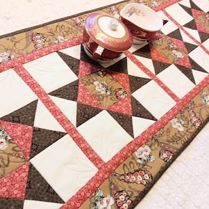 May include: A quilted table runner with a star pattern in white, brown, and pink. The runner features floral fabric accents and is topped with a red and white lidded sugar bowl and a matching creamer.