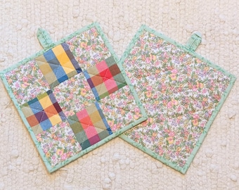 May include: Two square quilted pot holders. One features a patchwork design with plaid squares in yellow, blue, red, and green. The other has a floral pattern with pink, yellow, and green flowers. Both have a light green border and a hanging loop.