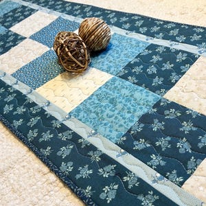 May include: A rectangular quilted table runner featuring a patchwork design of alternating squares in shades of teal, navy blue, and cream. The fabric squares display floral, star, and dotted patterns. Two round, woven rattan decorative spheres sit on top.